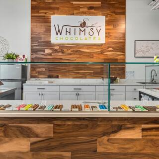 A cozy chocolate shop features a wooden counter, glass display of colorful chocolates, and a welcoming sign with a whimsical design.