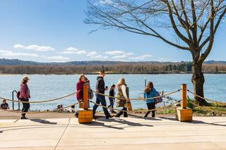A group of people strolls along a walkway by the water under a clear blue sky, surrounded by trees and nature.