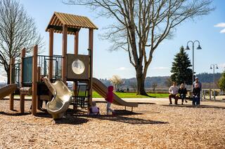 A playground features slides and play structures, while children explore nearby. Parents relax on a bench under clear skies and trees.