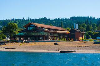 A large, modern building with a wooden structure sits near a sandy shore, surrounded by trees and water under a clear blue sky.