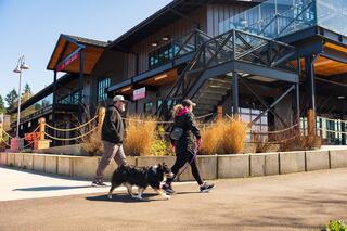 A couple strolls with a dog along a path in front of a modern building, enjoying a sunny day with greenery around.