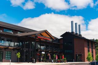 A modern, two-story building features a blend of brick and wood, with large windows and smokestacks, under a bright blue sky.