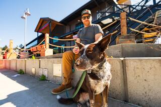 A man in a hat sits casually with an Australian shepherd on a leash, enjoying a sunny day near a modern building.