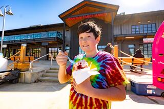 A smiling child in a tie-dye shirt enjoys a colorful dessert at a market, with sunny weather and people in the background.