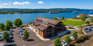 A waterfront scene features a modern building, green lawns, and nearby parked cars, with ships on the river and lush hills in the background.