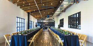 A spacious, elegantly set dining area featuring long tables adorned with dark linens and greenery, illuminated by soft overhead lights.