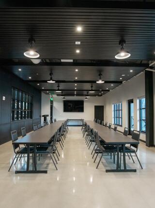 A modern conference room features a long table with chairs, sleek black ceiling lights, and large windows, creating a bright, professional atmosphere.