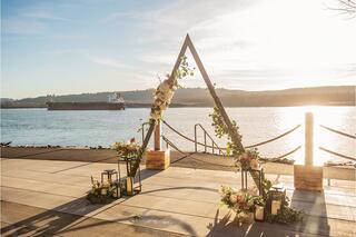 A floral-decorated triangular arch stands by the water, framed by a sunset glow, with a large ship passing in the background.